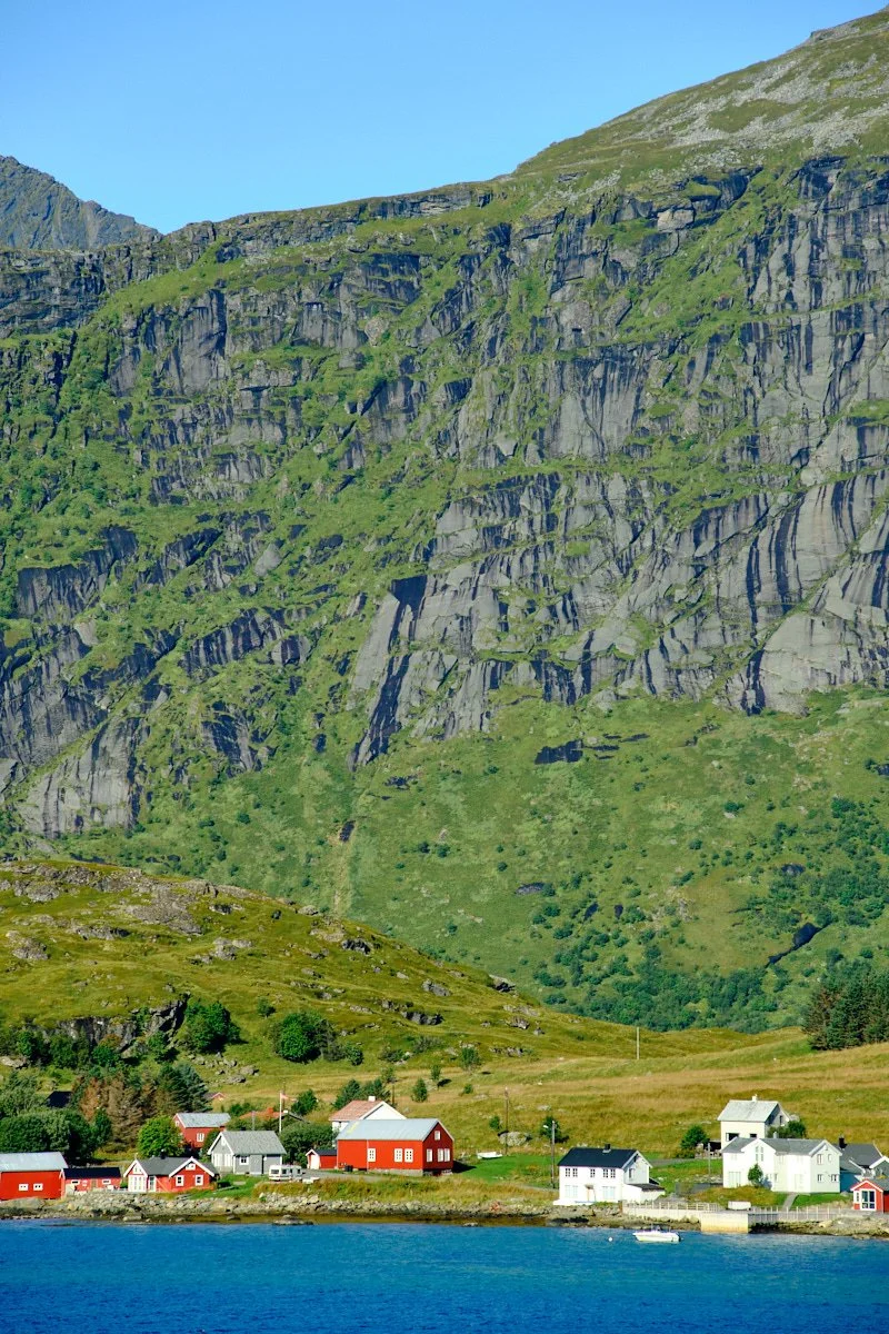Colorful houses by the water with a large green mountain in the background under a clear blue sky.