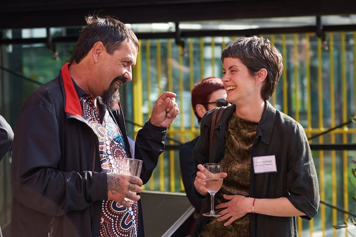 Two people at an outdoor event, smiling and talking. The man on the left has a beard, tattoos, and is holding a glass, while the woman on the right has short hair, a name tag, and a glass of water.