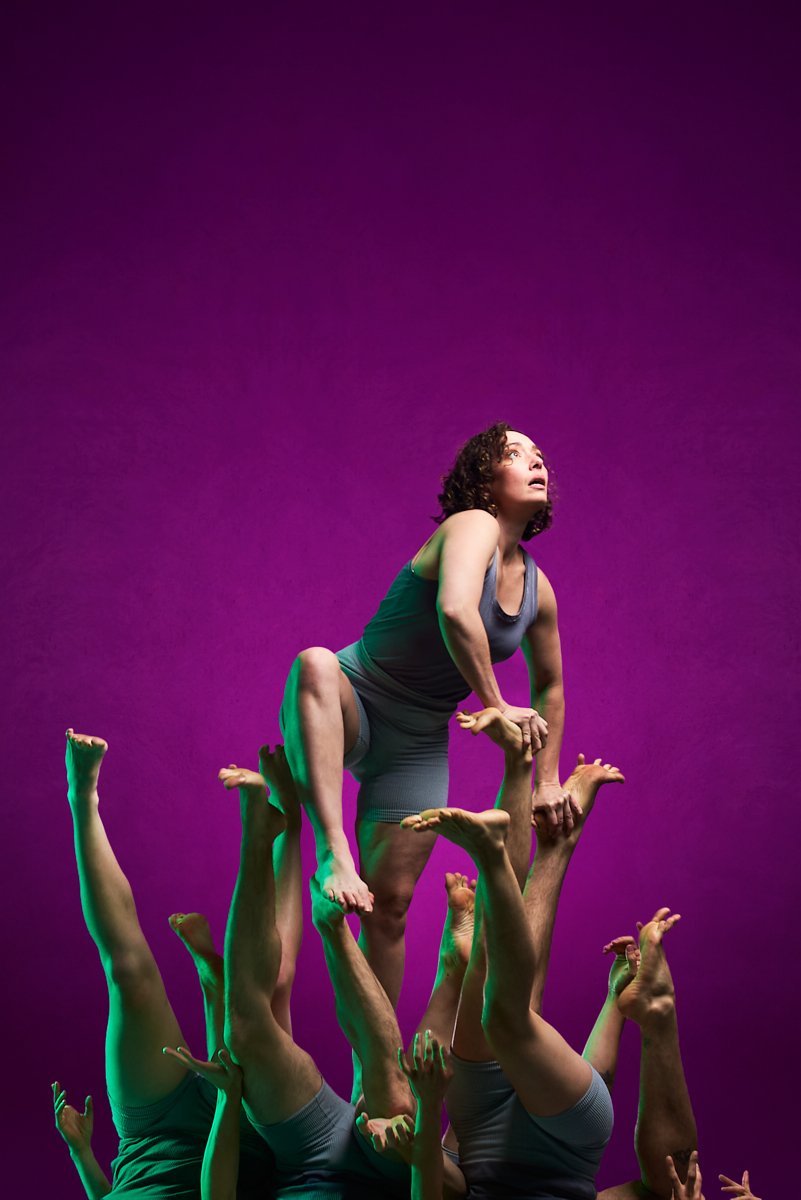 A woman is balanced on top of several people's legs and arms, performing a human pyramid against a purple background. She is looking up with an intense expression, wearing a gray tank top and shorts.