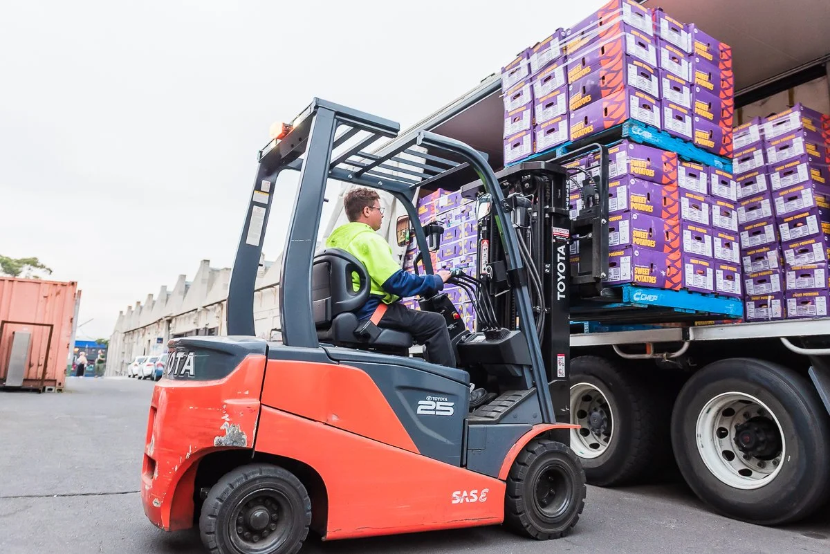 A man operating a red Toyota forklift is moving purple boxes labeled "Sweet Potatoes" onto a truck at an outdoor loading area.