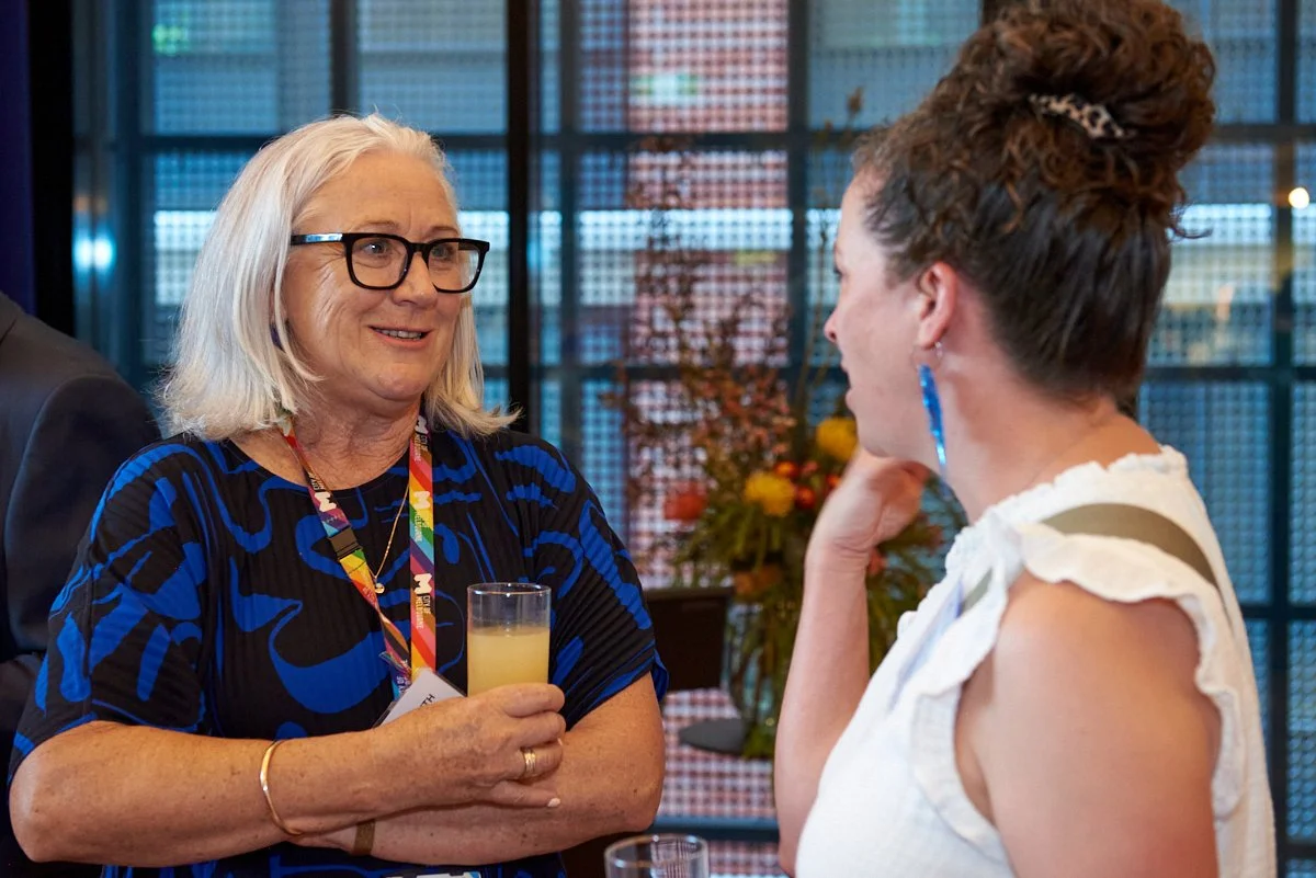 Two women having a conversation at a social event, one with gray hair and glasses holding a drink, and the other with dark, curly hair in a bun wearing a white top, in a modern indoor setting with flowers in the background.