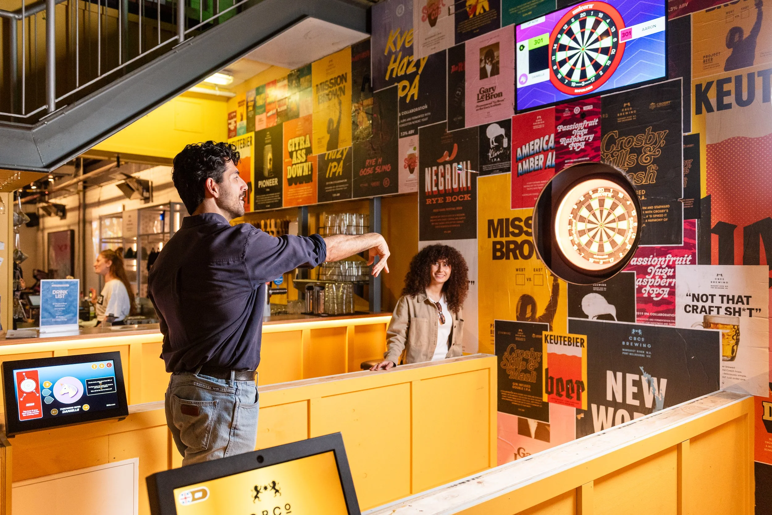A man reaches out to a dartboard on a colorful wall decorated with numerous posters at a bar. A woman stands behind a yellow counter, smiling, and there are other patrons and a digital device at the counter.