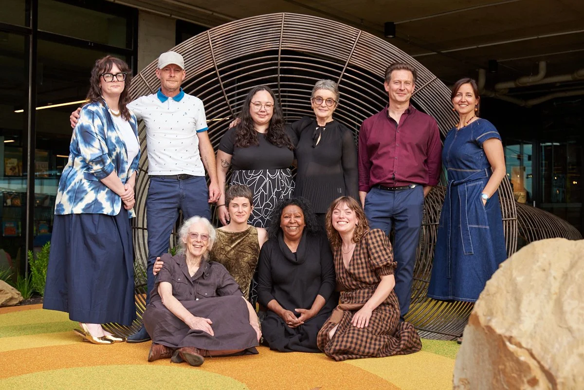 Ten diverse people posing for a group photo indoors with a modern, curved wooden art installation in the background. They are smiling and seated or standing on a colorful floor.