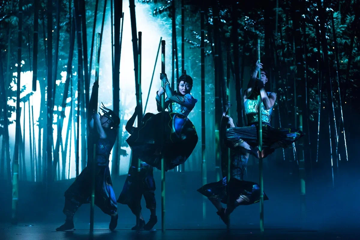 Four performers in traditional costumes performing a synchronized dance on stage with bamboo poles in a bamboo forest setting, illuminated with blue lighting.