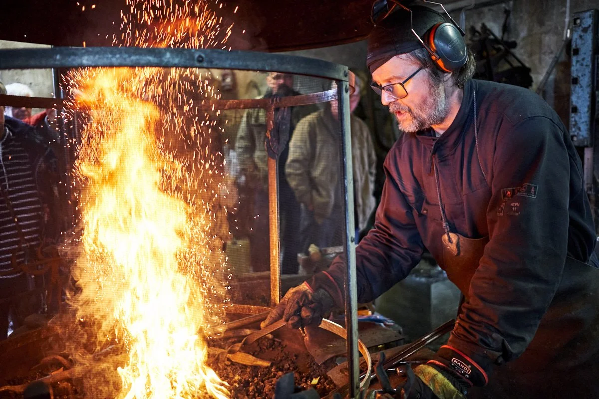 Blacksmith working with fire in a forge, sparks flying as he shapes metal, wearing safety glasses, gloves, and headphones, with a group of people observing in the background.