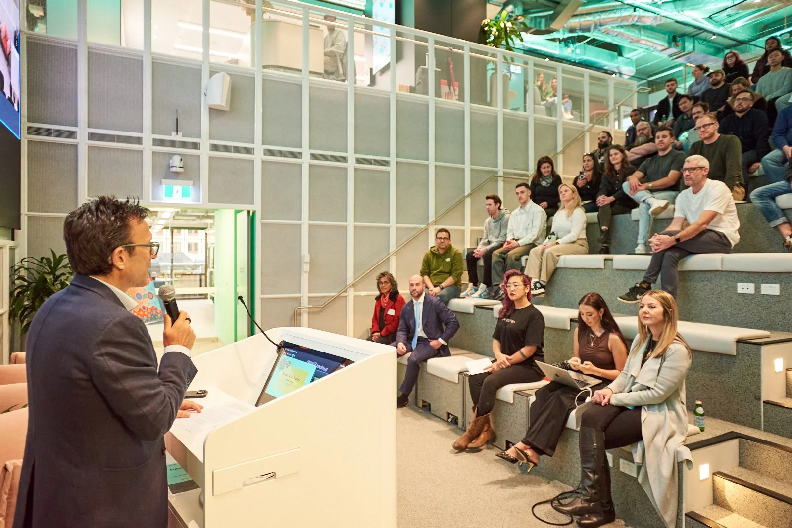 A man in a suit holding a microphone and speaking at a white podium during a presentation in a conference room. An audience is seated on tiered bleacher seating listening attentively.
