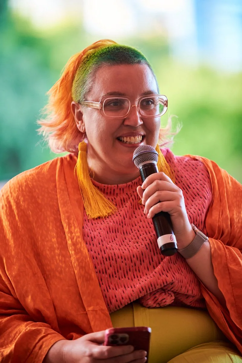 A woman with multicolored hair, wearing glasses, holding a microphone, and looking happy in a brightly lit indoor setting.