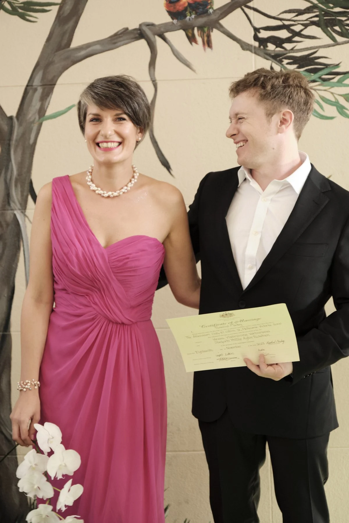 A smiling woman in a pink dress and a pearl necklace standing next to a man in a black suit, holding a marriage certificate, with a decorative tree and a parrot in the background.