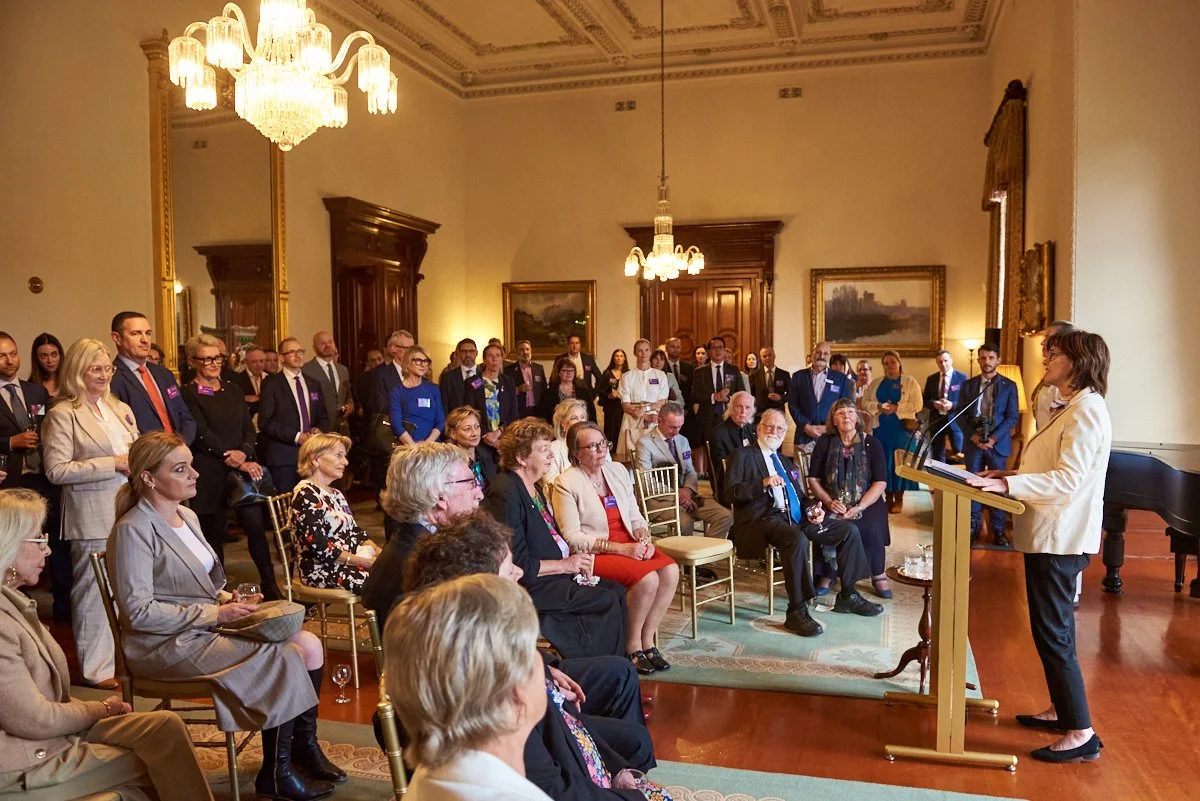 A woman is giving a speech at a podium in a formal, historic room with chandeliers, paintings, and wood paneling. An audience of men and women, some seated and others standing, listen attentively.