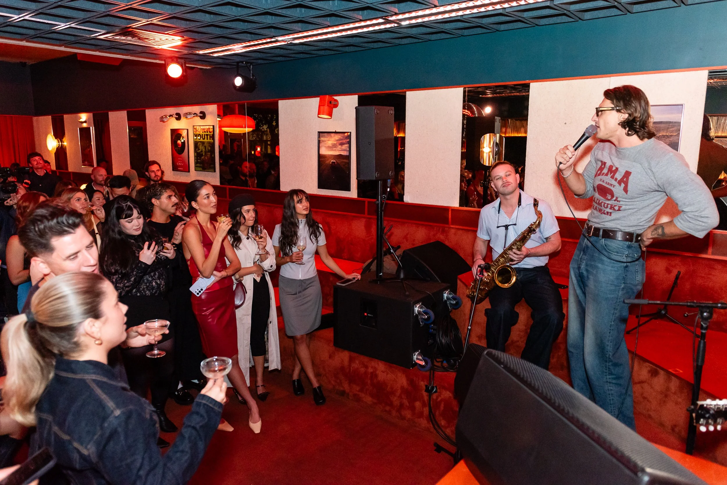 A woman with glasses singing into a microphone during a live performance at a bar or club. A man with a saxophone is seated beside her. The audience, mostly women, are watching and holding drinks. The room has red and blue lighting, and framed pictur