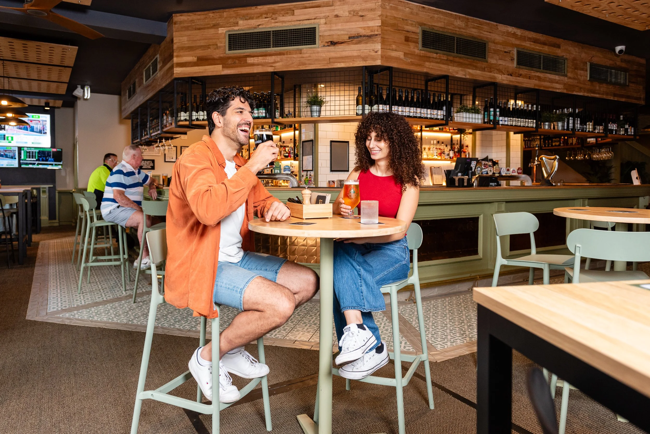 A man and woman are sitting at a round table in a restaurant, each holding a drink. The man is wearing a white T-shirt and orange jacket with denim shorts, and the woman is wearing a red sleeveless top and blue jeans. They are smiling and looking at 