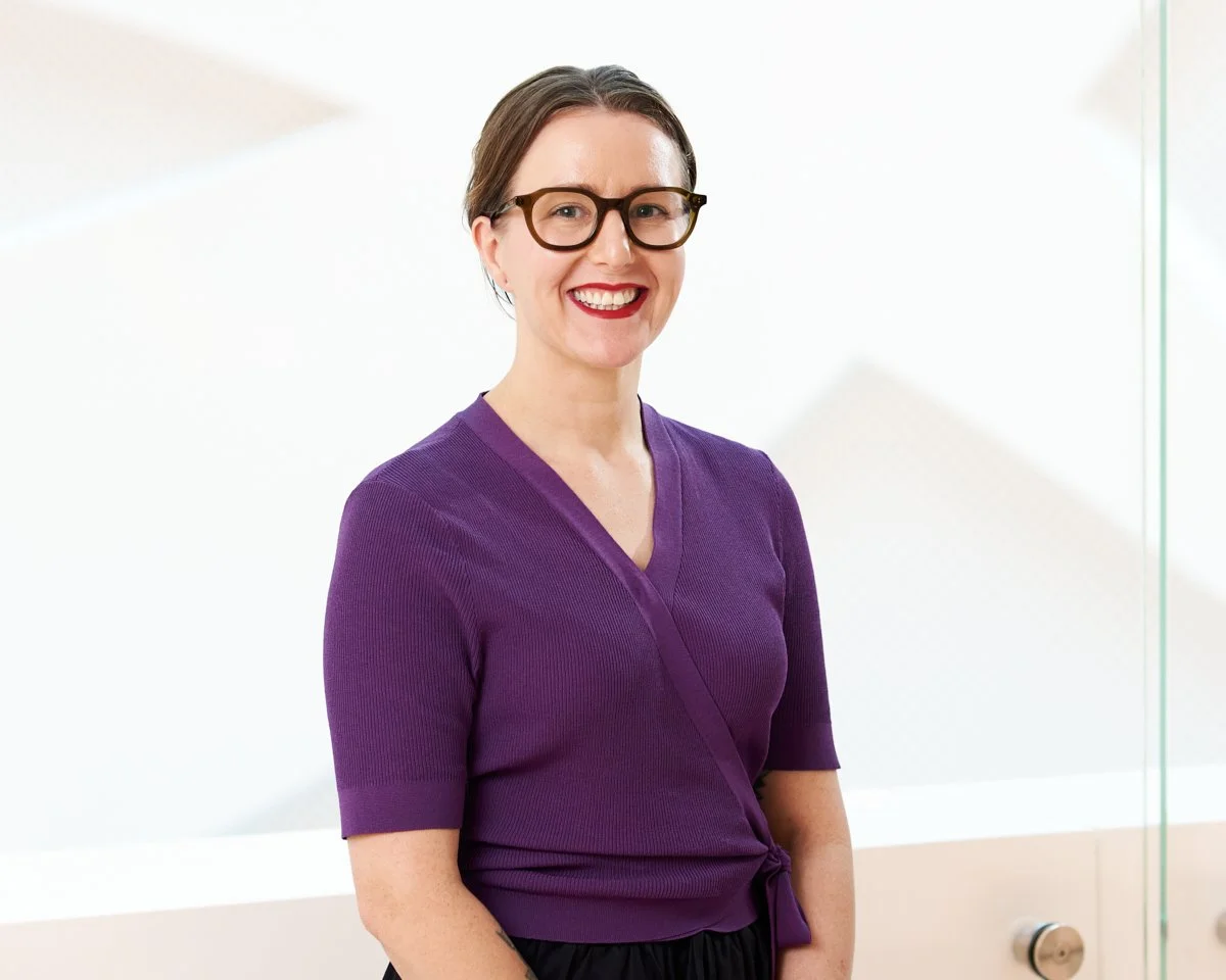 A woman with short brown hair, wearing glasses, a purple top, and black pants, standing and smiling in front of a light-colored background.
