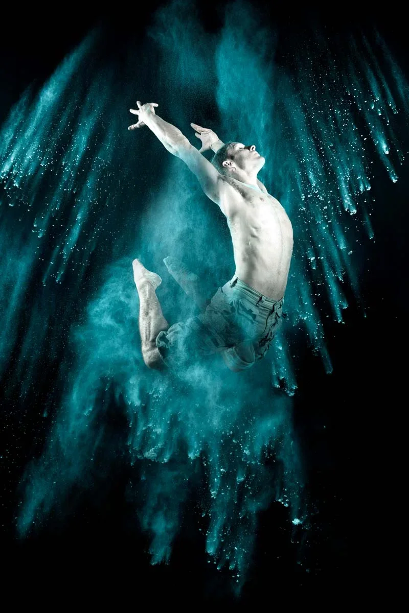 A man submerged underwater is captured mid-air, with water surrounding him in a dynamic splash, performing a dance or leap with arms extended upwards and head tilted back.
