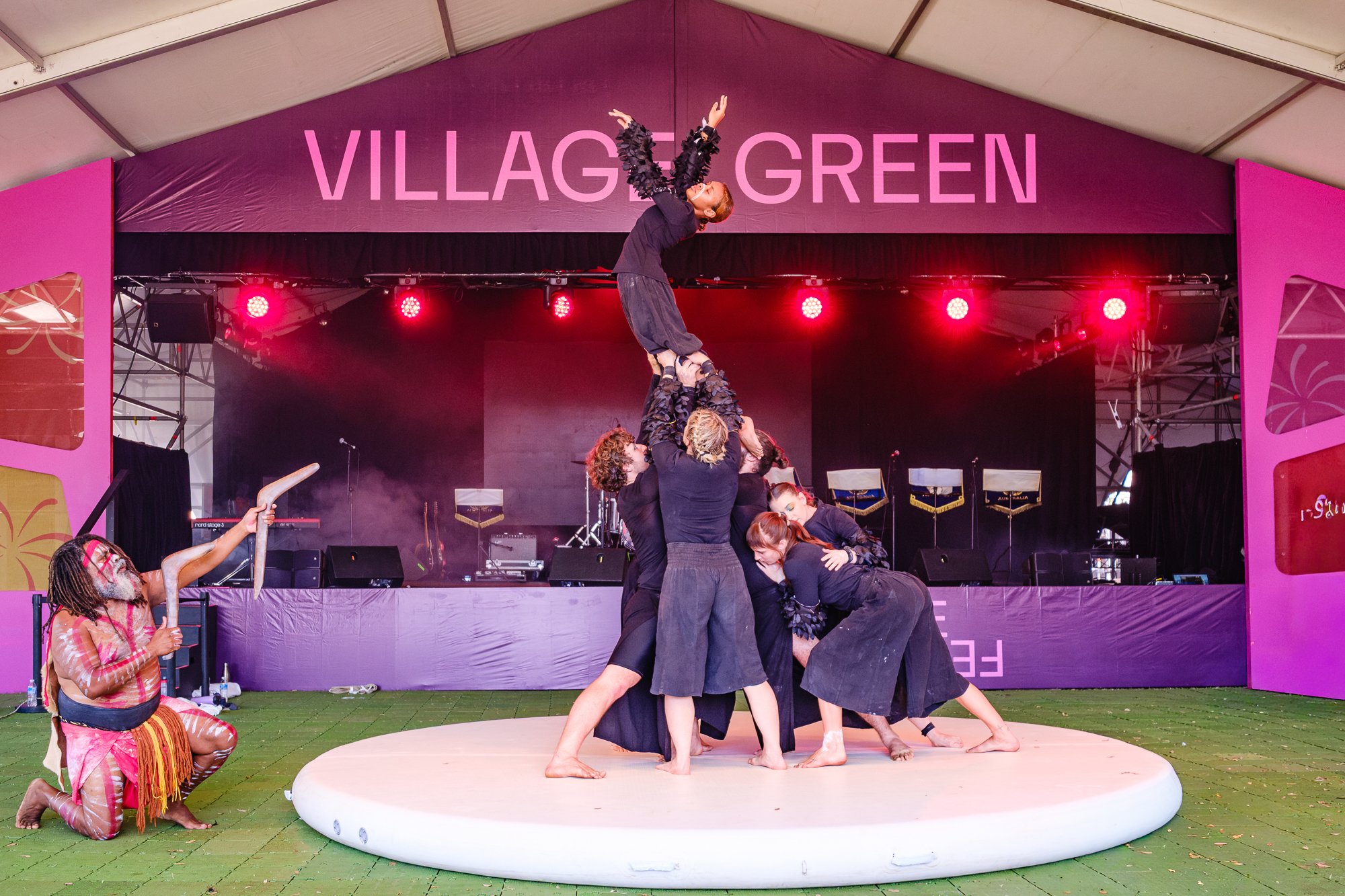 Group of performers in theatrical costumes performing a balancing act on stage under a large purple sign that reads 'VILLAGE GREEN'. The performers are supporting a woman in a black dress with ruffled sleeves, who is standing on their hands and leani