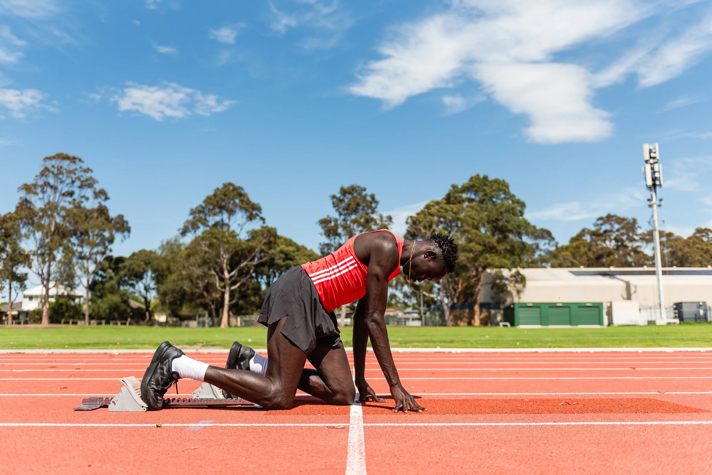 A male athlete in a red tank top and black shorts kneels on a red running track, preparing for a race with his head down and one hand on the ground. The setting is an outdoor track field with green grass, trees, and a blue sky with some clouds.