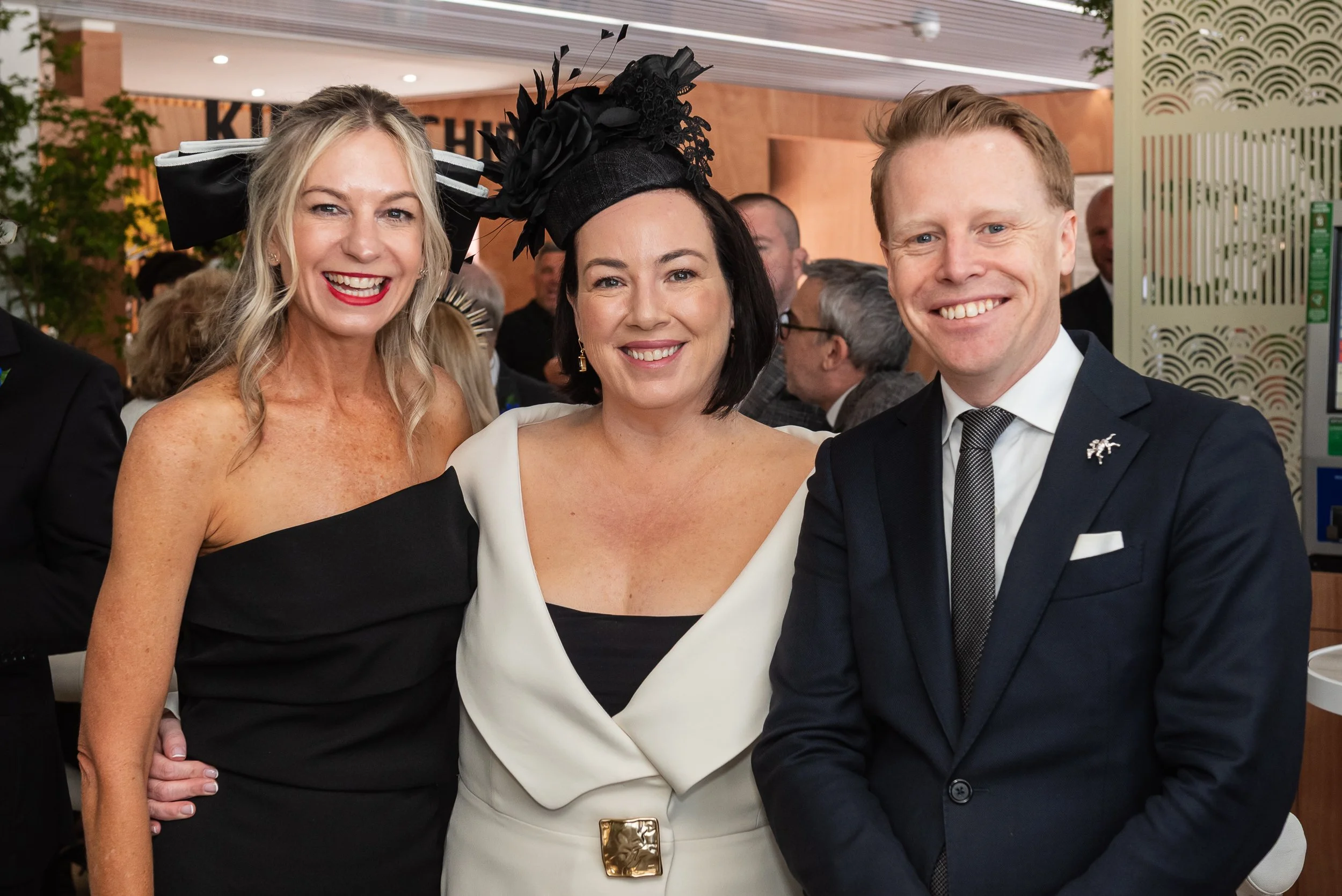 Three smiling people posing together at a formal event, with two women on the left and center and a man on the right, all dressed in elegant attire.