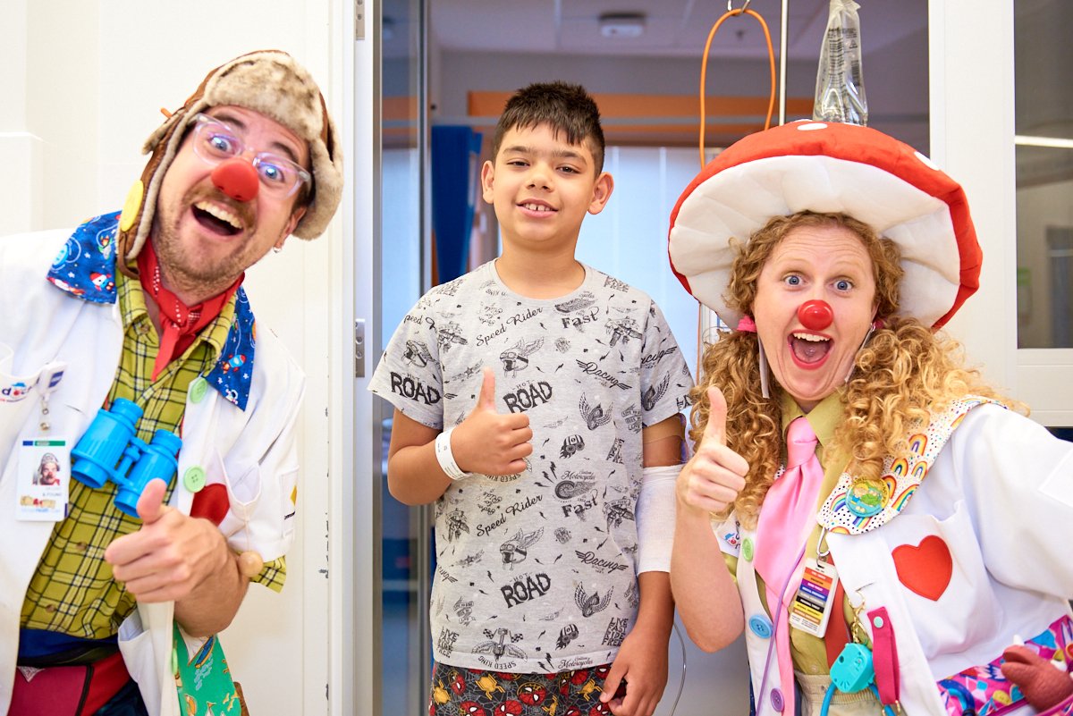 A young boy in hospital gown standing with two clowns dressed as medical professionals in a hospital corridor. Both clowns are smiling and giving thumbs-up gestures, wearing colorful costumes, red noses, and clown accessories.