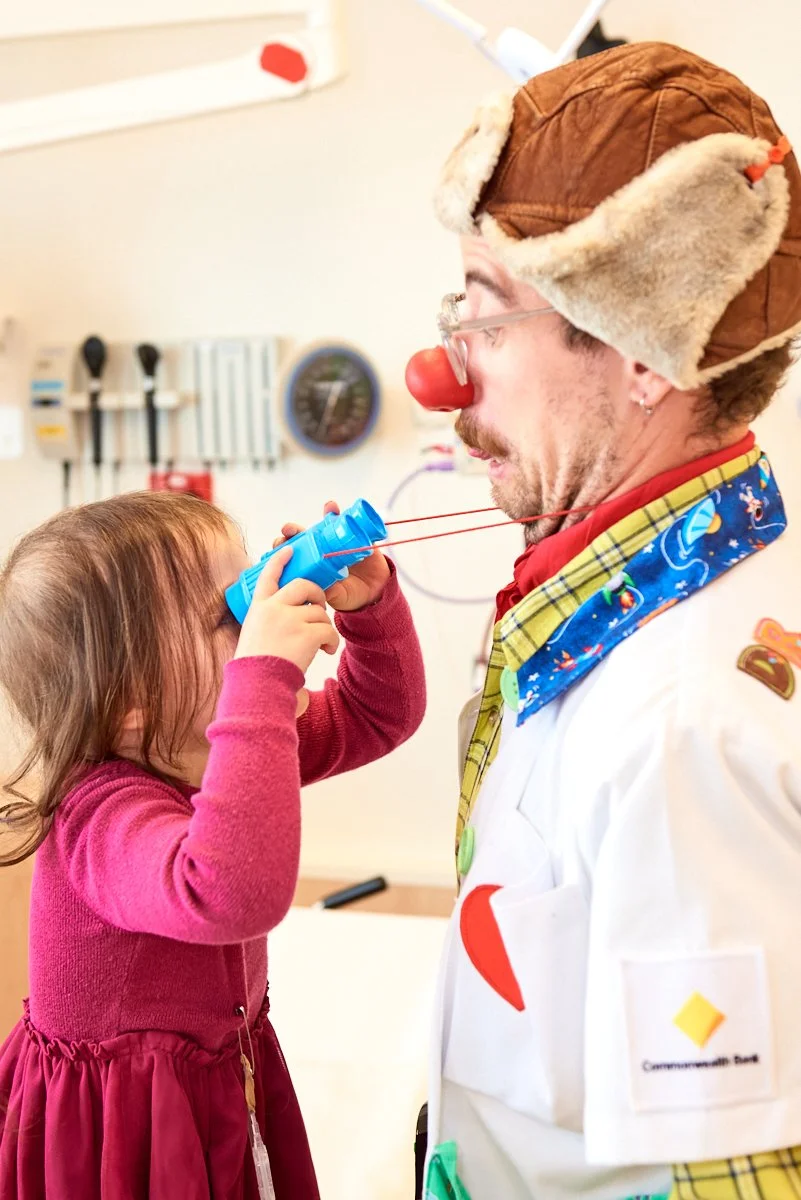 A young girl in a pink dress blowing a party blow horn with a clown nose on a man dressed as a clown or doctor with a brown hat and colorful scarf in a medical room.