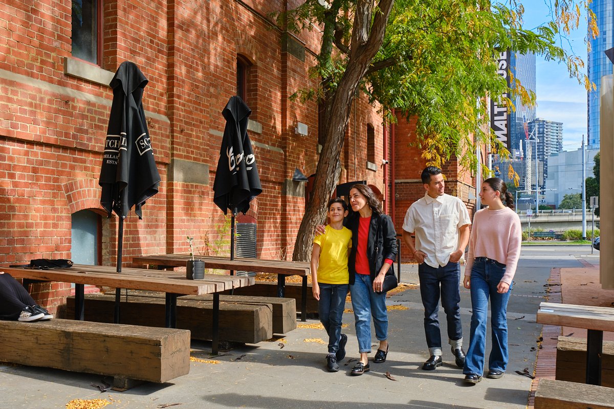 Four people walking on a city sidewalk near a brick building with outdoor seating and umbrellas, trees, and high-rise buildings in the background.