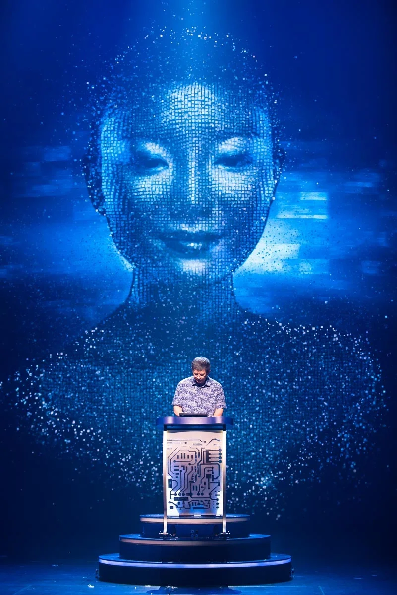 A man stands at a podium with a digital circuit pattern, with a large digital portrait of a smiling woman's face projected behind him, both in a blue-themed digital art setting.