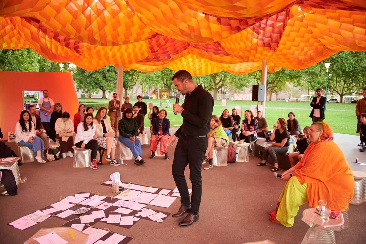 A man holding a microphone speaks to an audience seated outdoors under a large orange geometric canopy. Papers and a cup are on the ground in front of him. The audience appears engaged, with some smiling and others listening attentively. Trees and gr