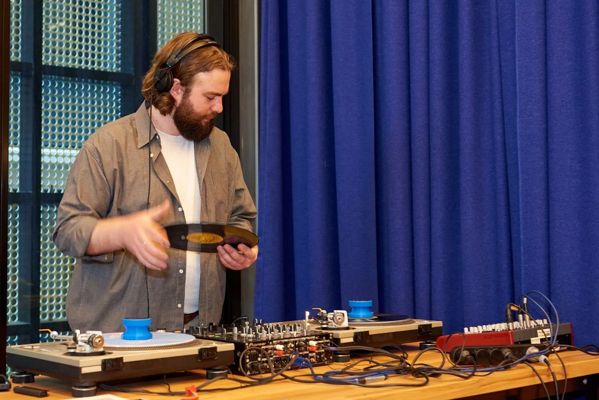 A man with long hair and a beard wearing headphones, holding a vinyl record, standing behind DJ equipment on a table with blue curtain backdrop.