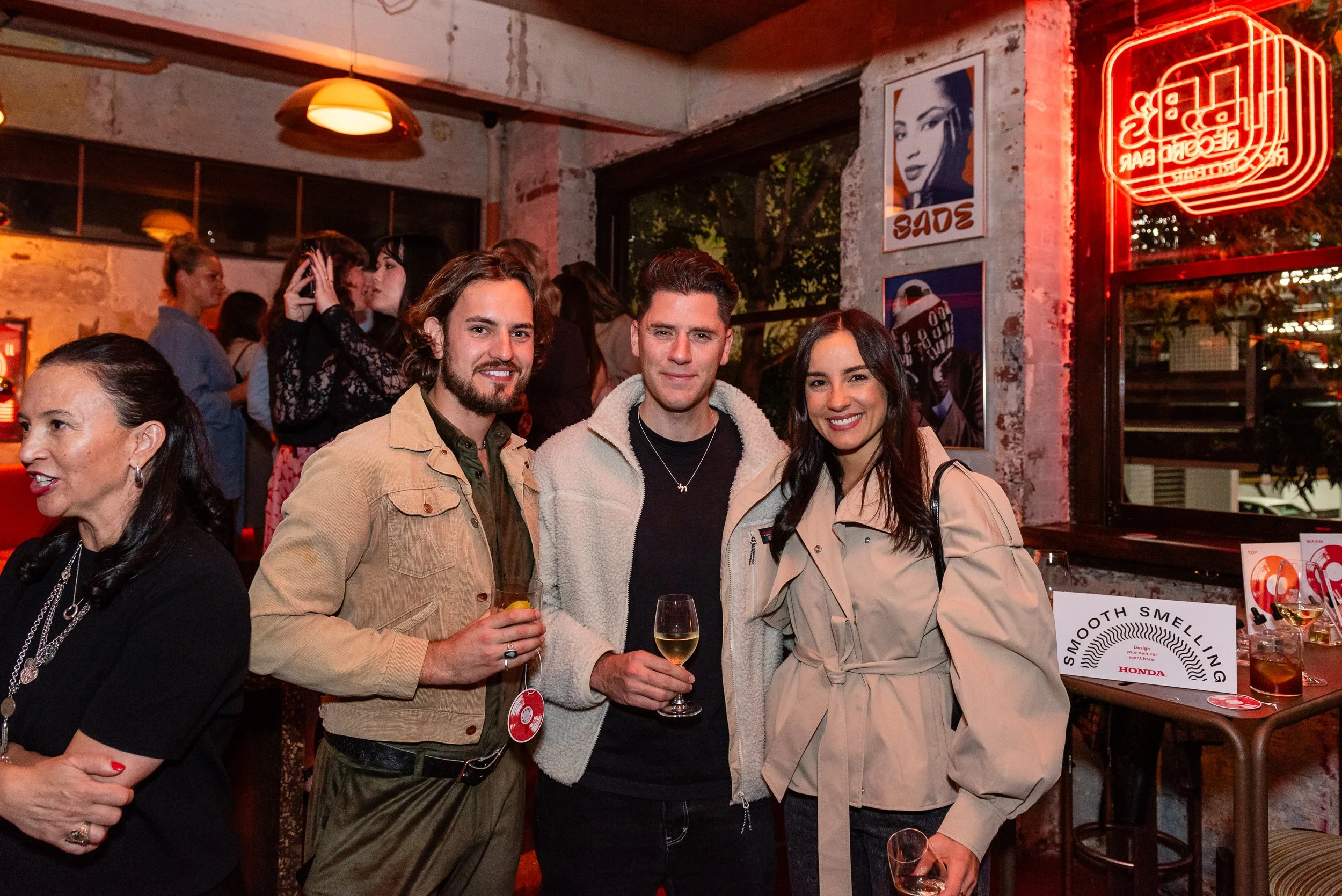 Three young adults standing together at a social event, smiling and holding drinks, with a crowd in the background inside a lively bar or restaurant.