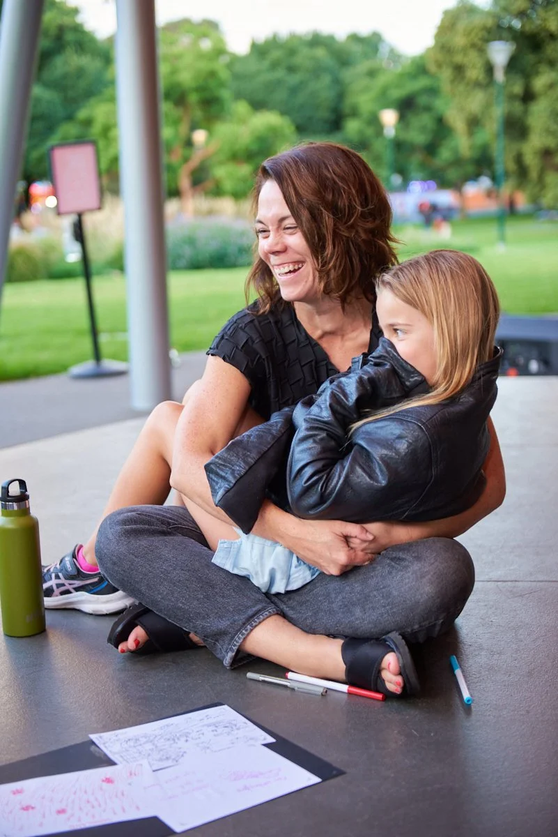 A woman and a young girl sitting outside on a concrete surface, with the woman hugging the girl. The woman is laughing and the girl appears shy. There are papers and markers on the ground, a green water bottle nearby, and a park with trees and lamppo