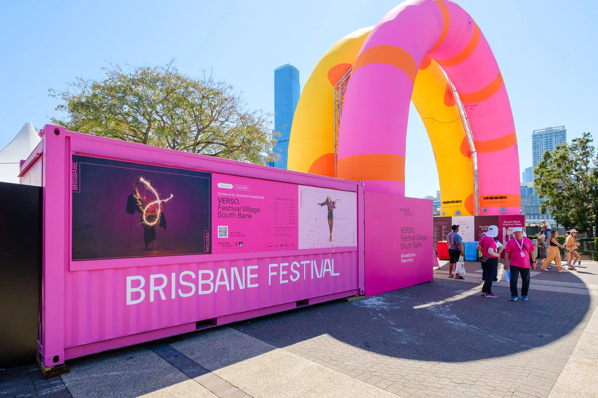 Pink temporary event structure with signs for the Brisbane Festival, featuring images and texts inviting people to attend, under a clear blue sky with city buildings and trees in the background.