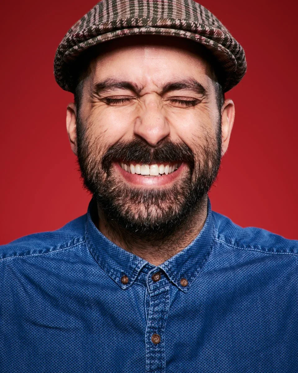 A man with a beard smiling with his eyes closed, wearing a flat cap and a blue button-up shirt, against a red background.