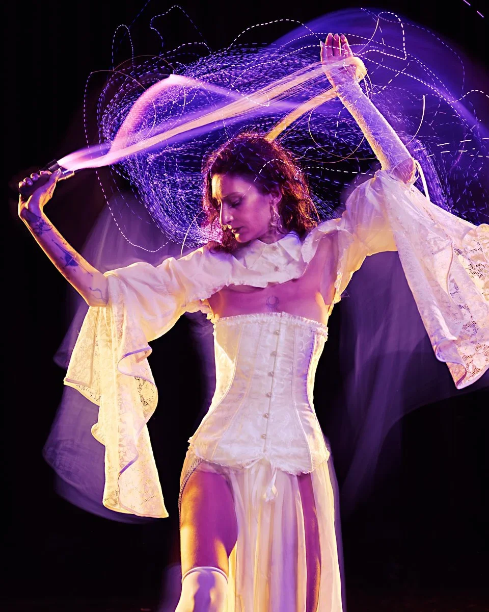 A woman in a cream-colored corset and flowing skirt performs with a violet laser sword, creating light trails in a dark setting.