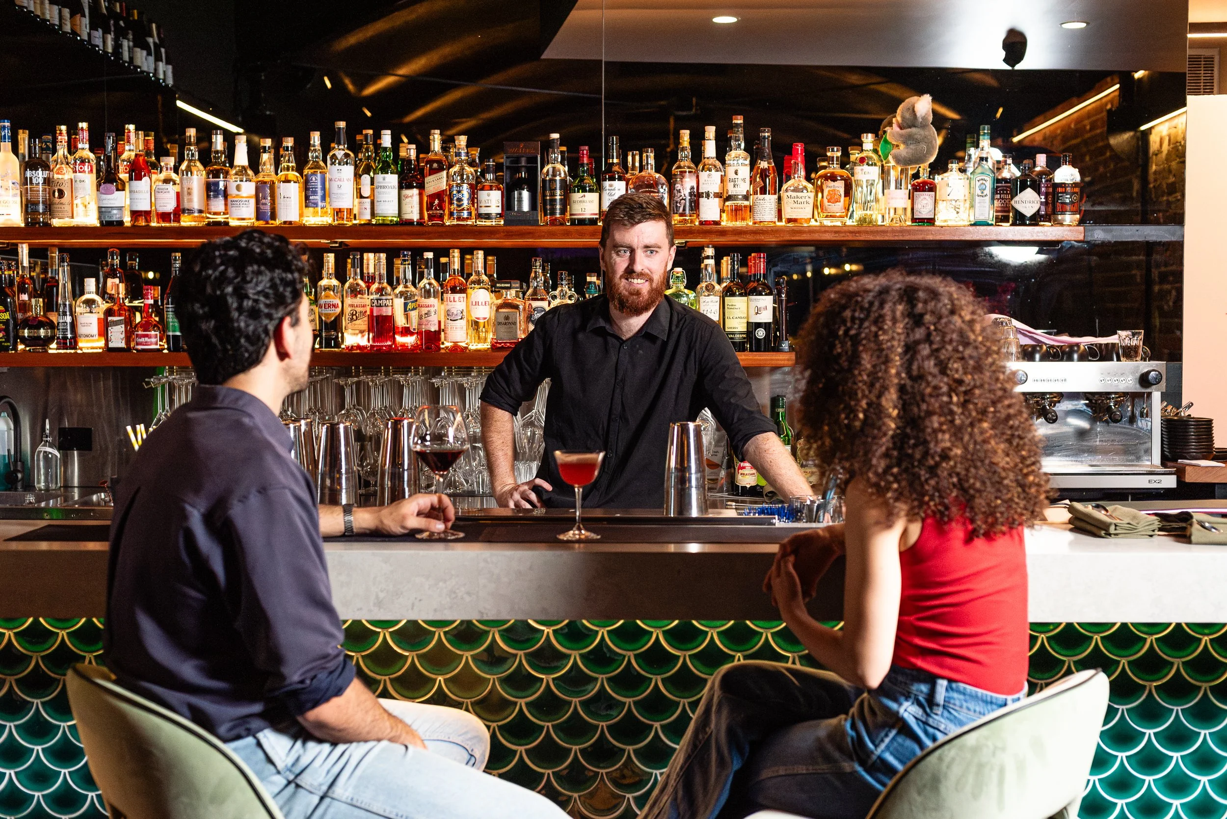 A bartender standing behind a bar counter, serving drinks to customers, with a background of various liquor bottles on shelves. Three patrons are visible, two men and a woman, engaged in conversation.