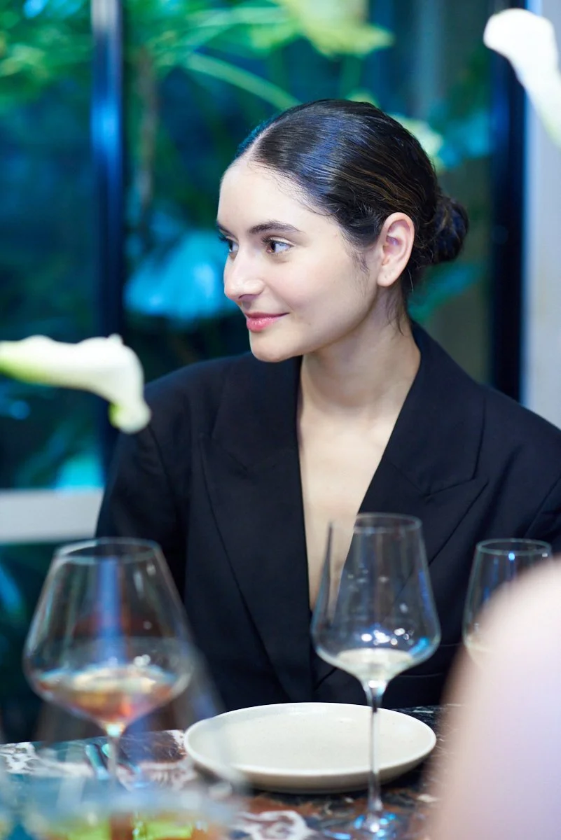 A young woman with dark hair tied back, wearing a black blazer, sitting at a dining table with empty glasses and a plate, smiling and looking to her left in a restaurant setting.