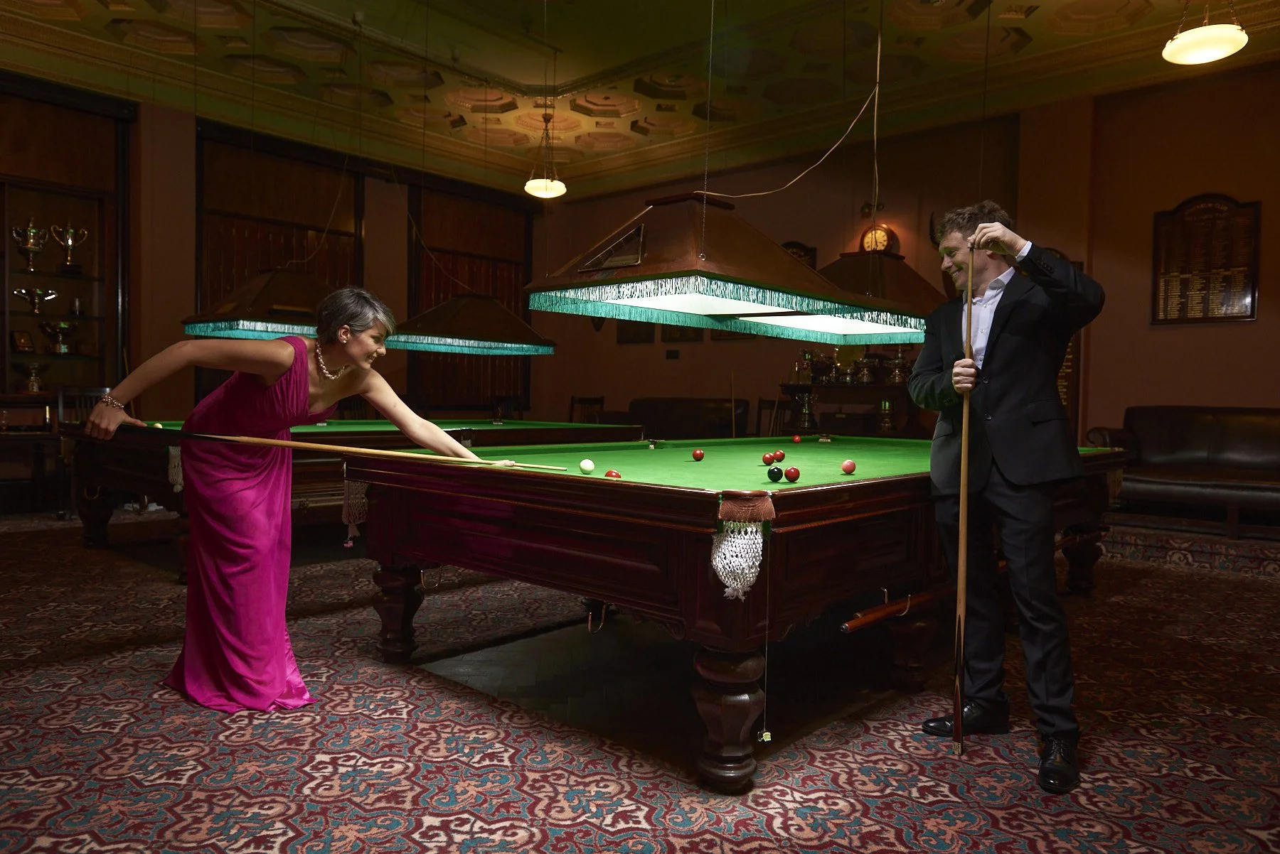 A woman in a pink dress and a man in a black suit playing pool in a dimly lit, vintage-style game room with wooden walls, patterned carpet, and hanging lamps.