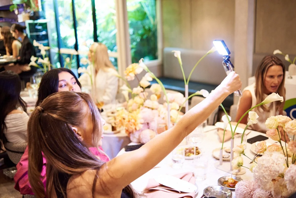 Women sitting at a decorated table with flowers, selfie-taking with a smartphone, in a bright room with large windows and lush greenery outside.