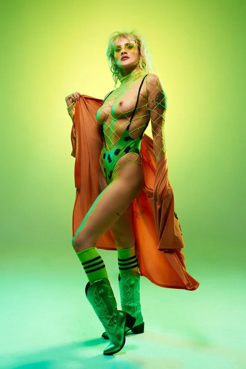 A woman wearing bright yellow sunglasses, fishnet green top, animal print bodysuit, orange trench coat, knee-high striped socks, and cowboy boots, posing against a green gradient background.