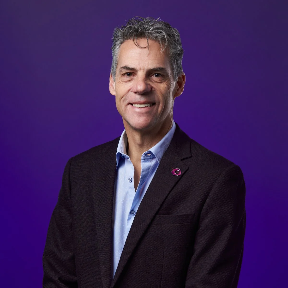 A middle-aged man in a dark blazer and blue shirt standing against a purple background.