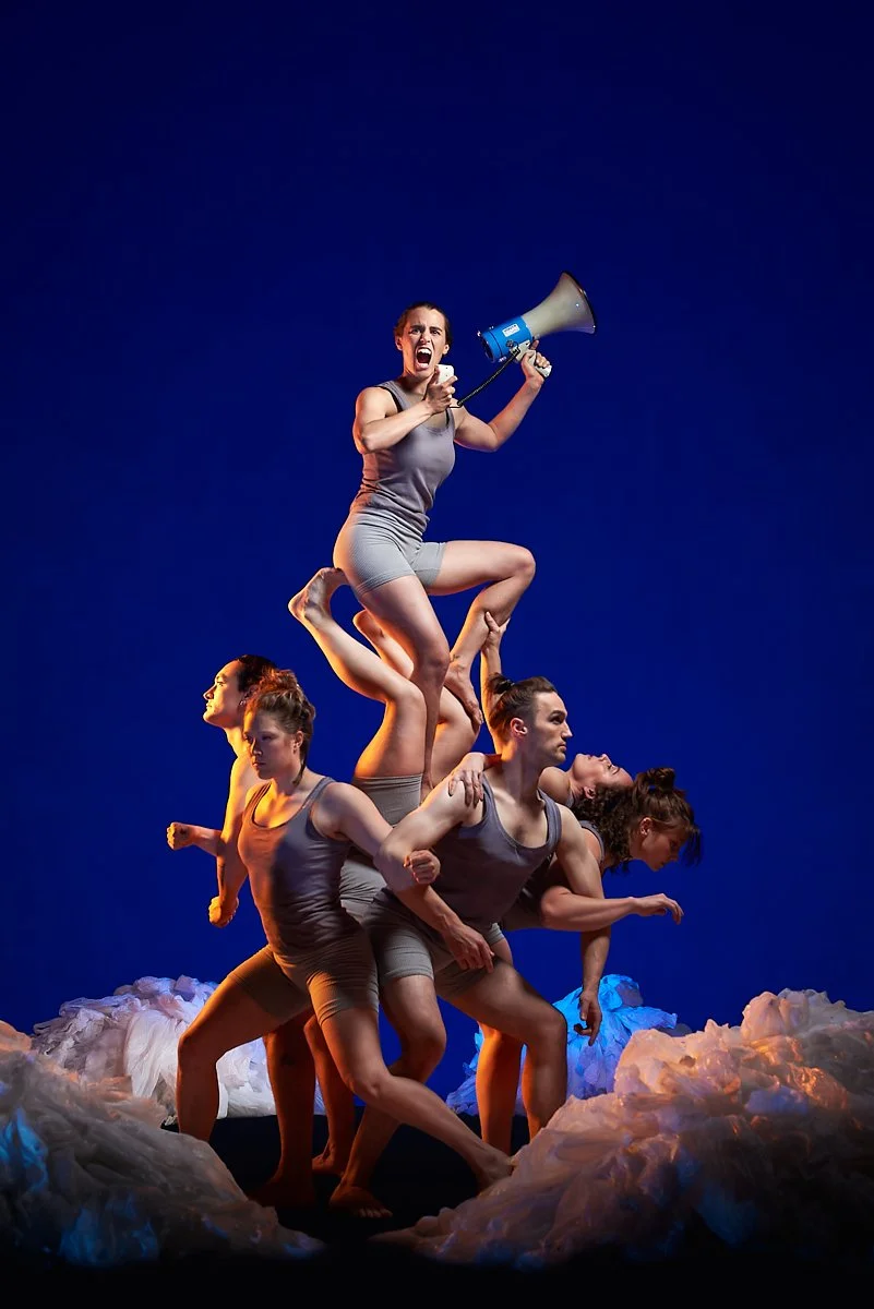 Five women in gray athletic clothing performing a human pyramid against a dark blue background, with the top woman holding a megaphone and shouting.