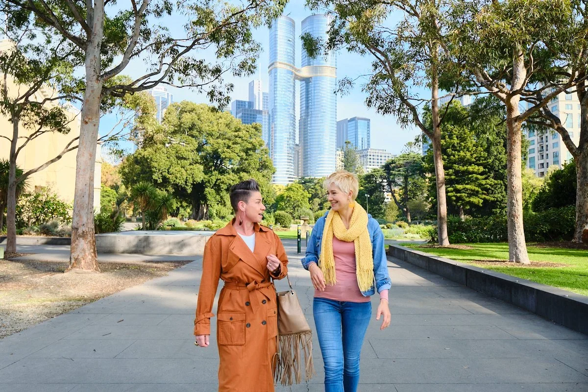 Two women walking and talking in a city park with tall modern buildings in the background.