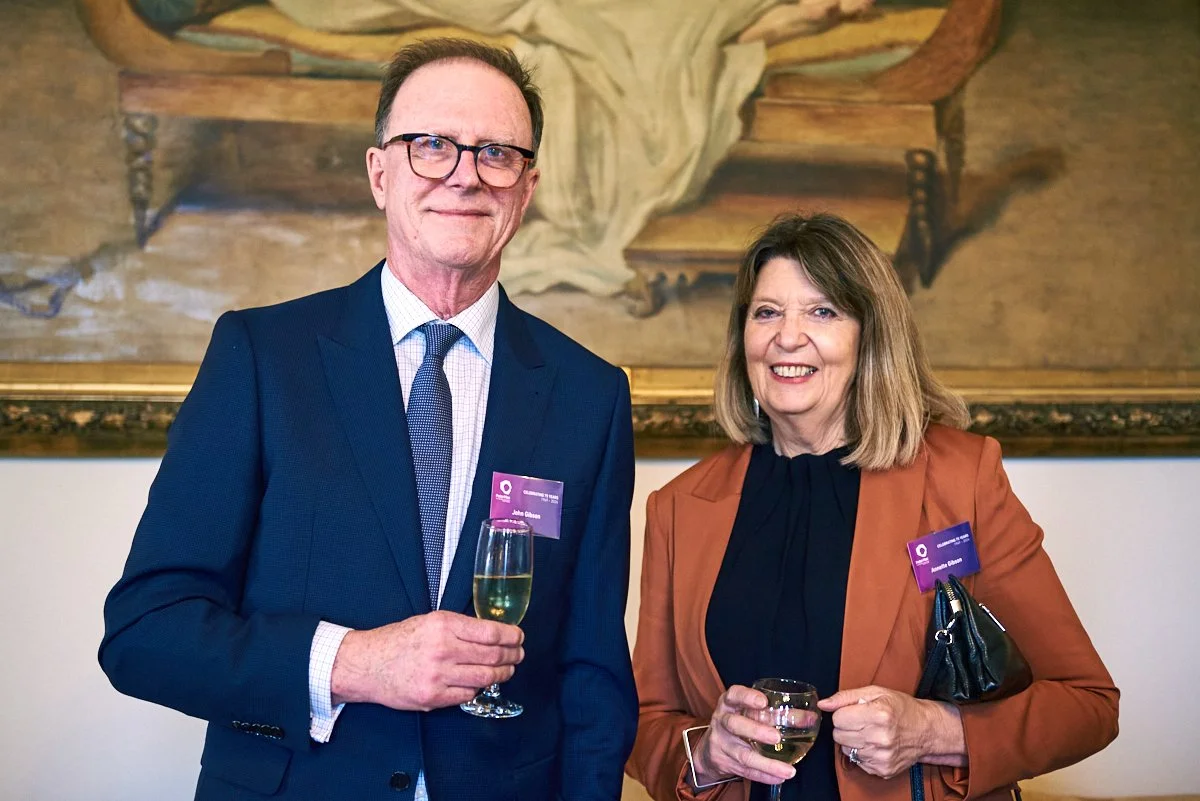 A man and woman in business attire attending a formal event, holding glasses of champagne, standing in front of a large painting.