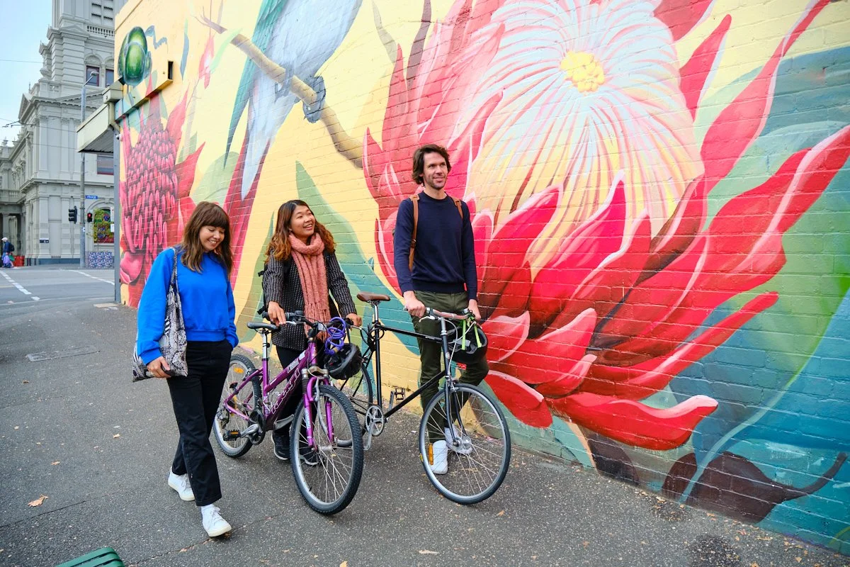 Three people with bicycles standing in front of a large, colorful floral mural on a brick wall in an urban area.