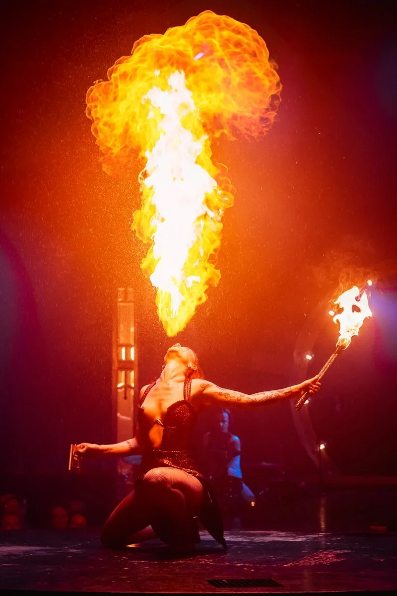 A female fire performer in a black outfit performing a fire-breathing act, producing a large plume of fire, during a nighttime show.