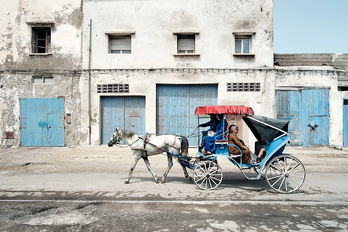 A horse-drawn carriage with a red canopy carrying two passengers and one driver, moving along a street in front of an old building with peeling white paint and blue doors.