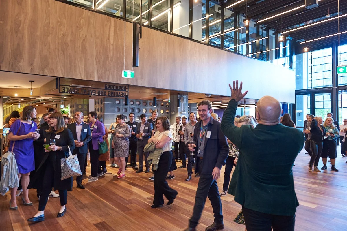 People gathering at a modern professional event or conference in a spacious, wood-finished interior with large glass windows and high ceilings. Some individuals are holding drinks, engaging in conversation, while a presenter with an outstretched hand