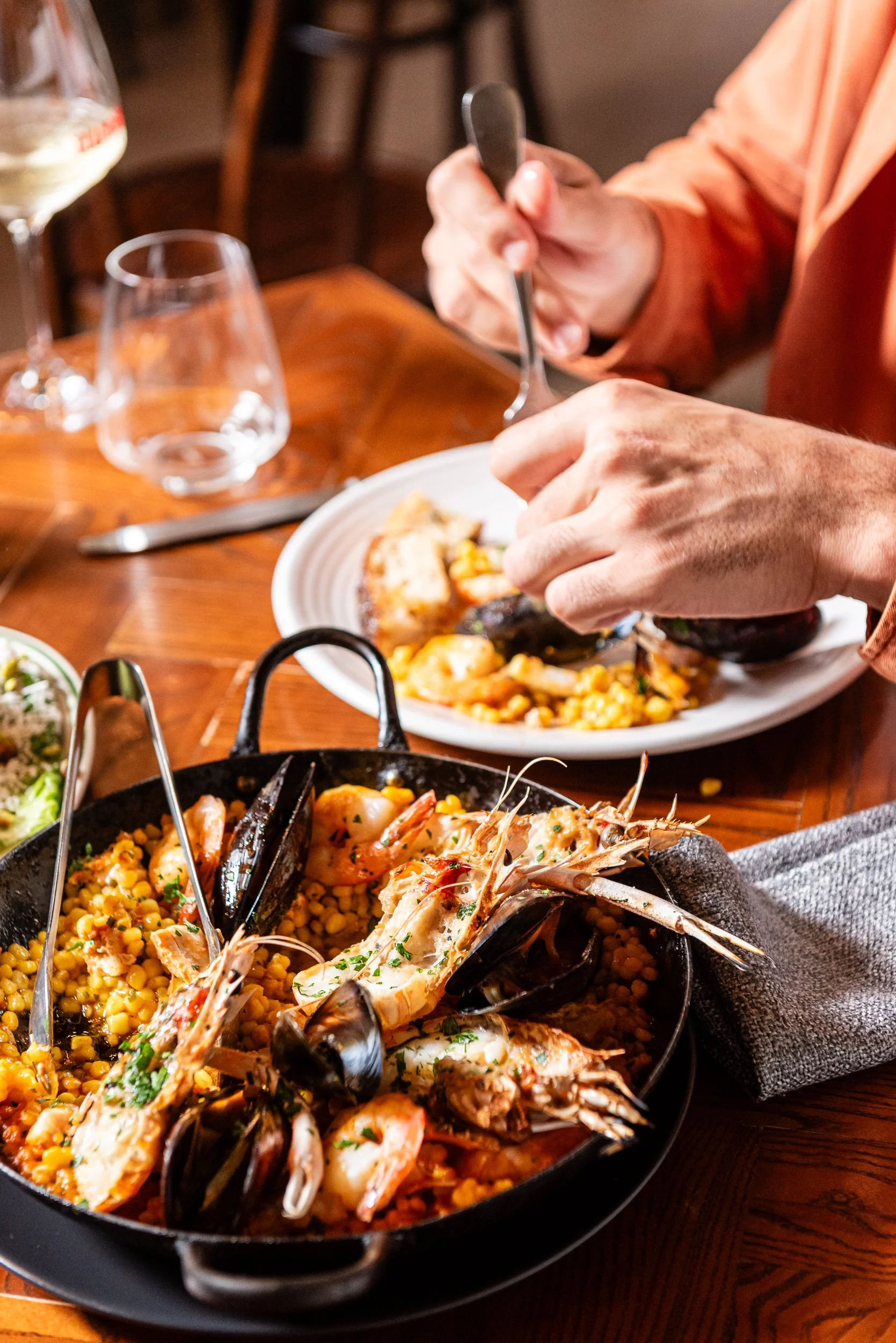 A close-up of a seafood dish with shrimp, lobster, and corn, garnished with herbs, served in a black skillet on a wooden table, with a person eating from a white plate in the background.