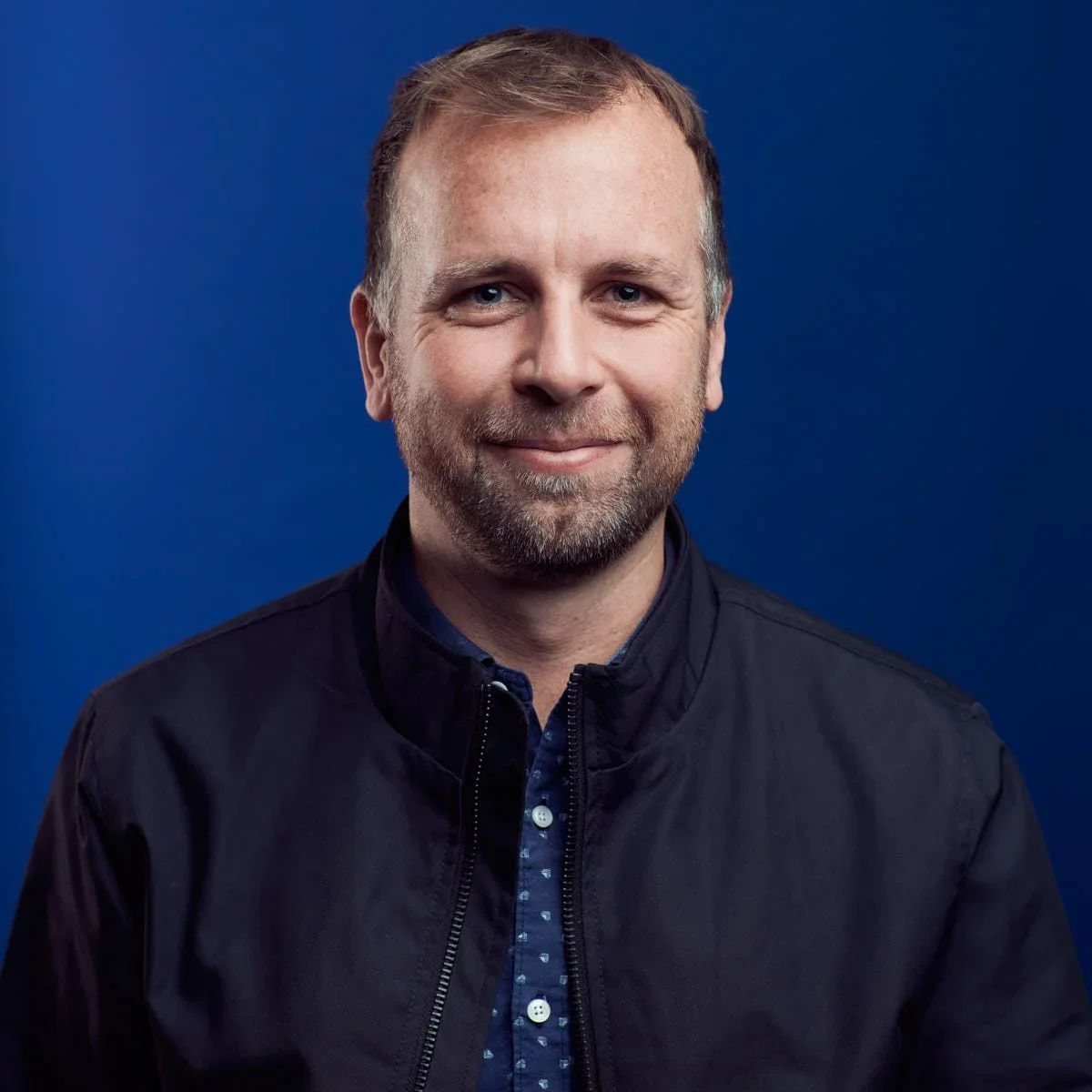 Portrait of a smiling man with light brown hair, blue eyes, and a beard, wearing a dark jacket and a collared shirt, against a blue background.