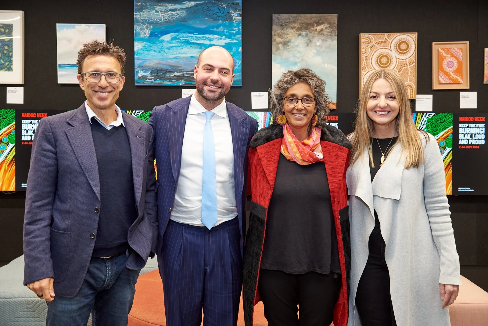Four people standing side by side, smiling in an art gallery or exhibit with colorful paintings and digital screens in the background.