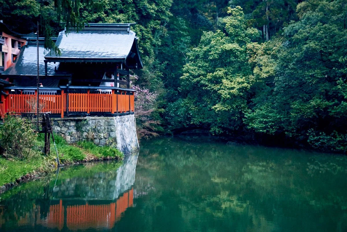 Traditional Japanese building with a slate roof and wooden balcony, situated on a stone foundation, overlooking a calm river surrounded by lush green trees.