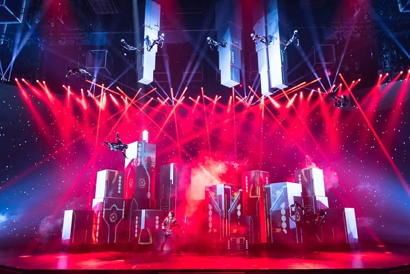 A concert stage with vibrant red and purple lighting, multiple vertical screens, and a performer in the center.
