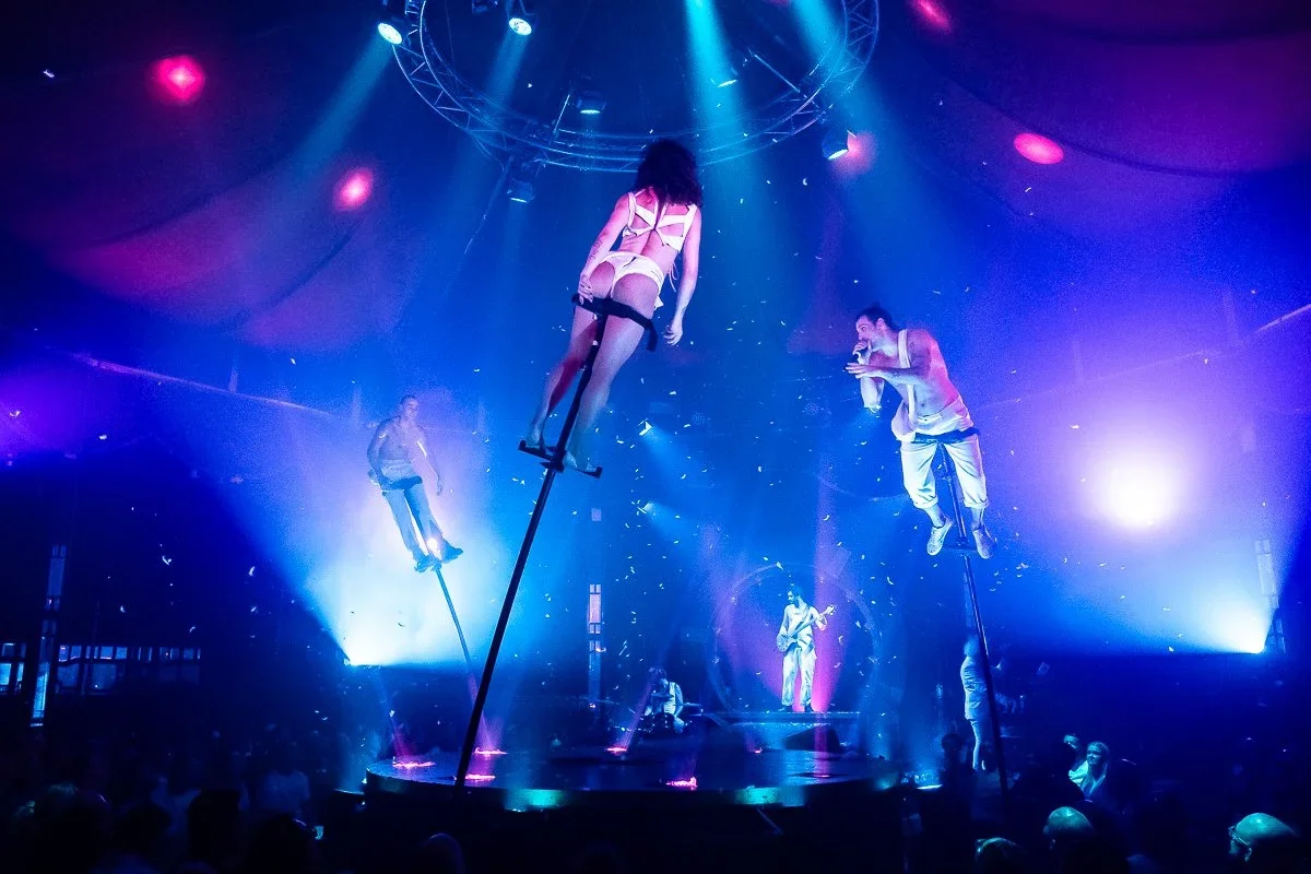 A circus or performance stage with performers on stilts and a singer, illuminated by vibrant blue and pink lights in a dark auditorium.